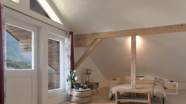 Un dormitorio con techo inclinado, vigas de madera, cama, mesita de noche, un jarrón con flores y vista a un bosque a través de una ventana.