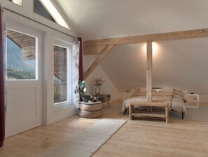 Ein Schlafzimmer mit schrägverdeckter Decke, Holzbalken, einem Bett, einem Nachttisch, einer Vase mit Blumen und Blick auf einen Wald durch ein Fenster.