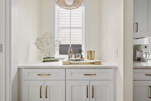 A kitchen with white cabinetry, gold handles, and a window with blinds.
