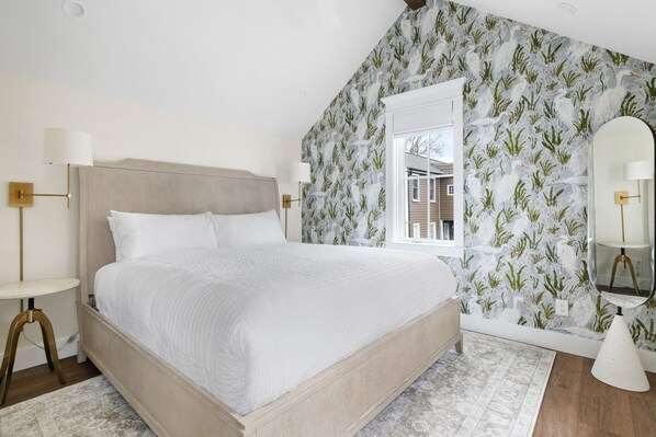 A bedroom with a large bed, a round side table, a mirror, and a window with a view of a house.