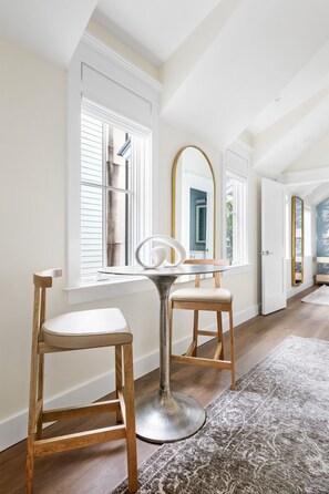 A room with a wooden chair and a table with a mirror. There are windows with blinds, a rug on the floor, and a door in the background. - The Pink Palm | Private Pool | Bach Trip Paradise (Charleston)