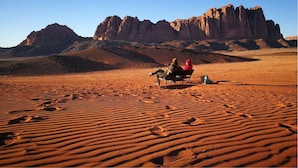 A desert landscape with a large rock formation in the background and two people sitting on a bench. - Wadi Rum Heritage (Wadi Rum)