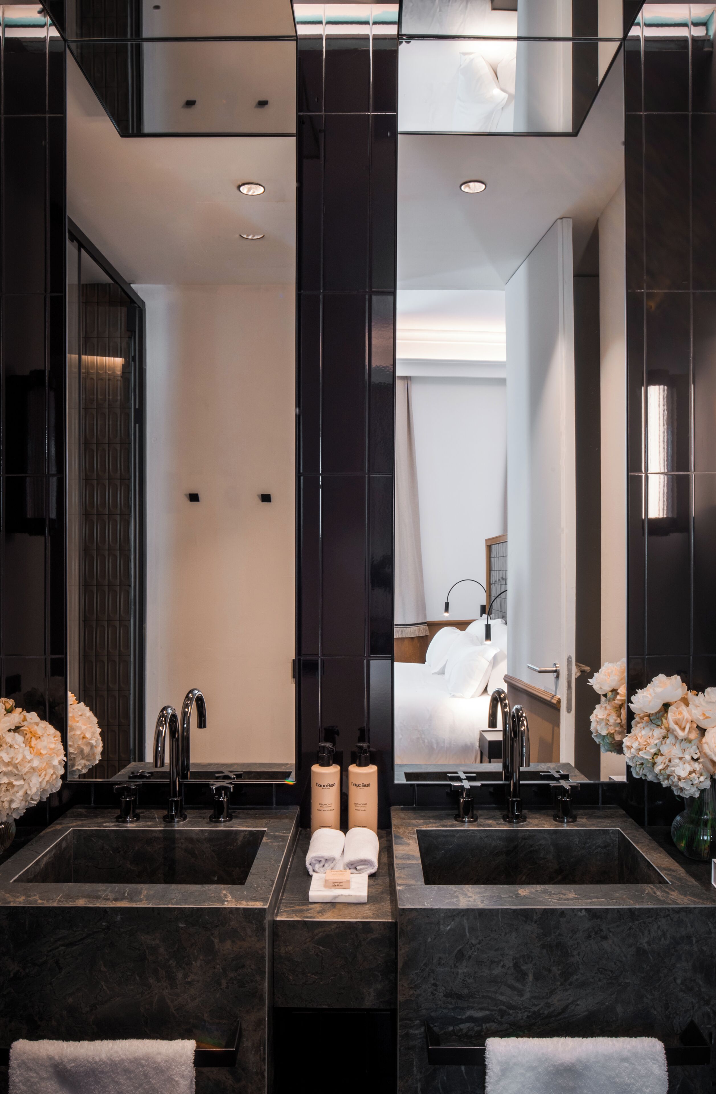 A modern bathroom with two sinks, a large mirror, and a bed visible in the reflection.