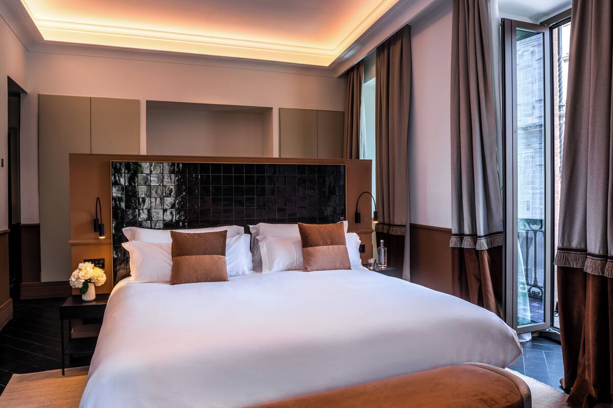 A large bed with a headboard, a bedside table with a lamp, and a window with curtains.