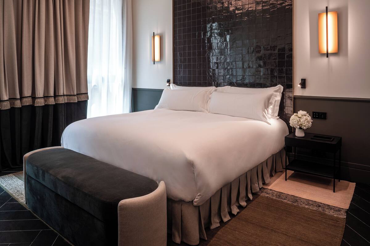 A large bed with a headboard, a bedside table with a vase of flowers, and a window with curtains.