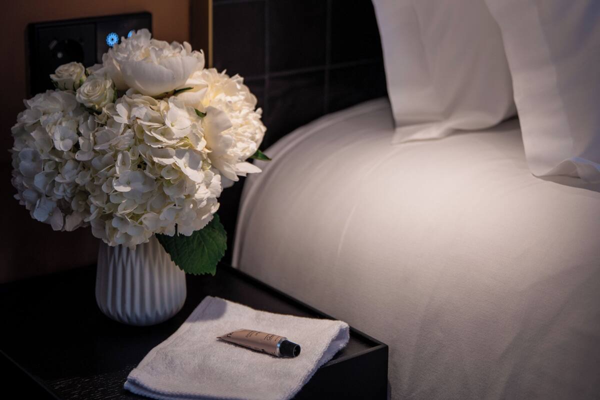 A hotel room with a bed, a nightstand with a vase of flowers, a lamp, and a remote control.