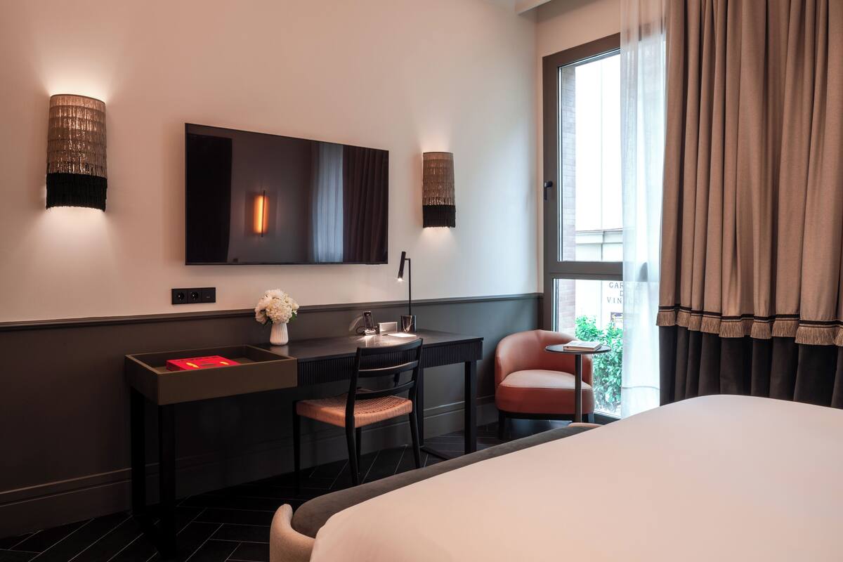 A hotel room with a bed, a desk with a chair, a TV mounted on the wall, and a window with curtains.