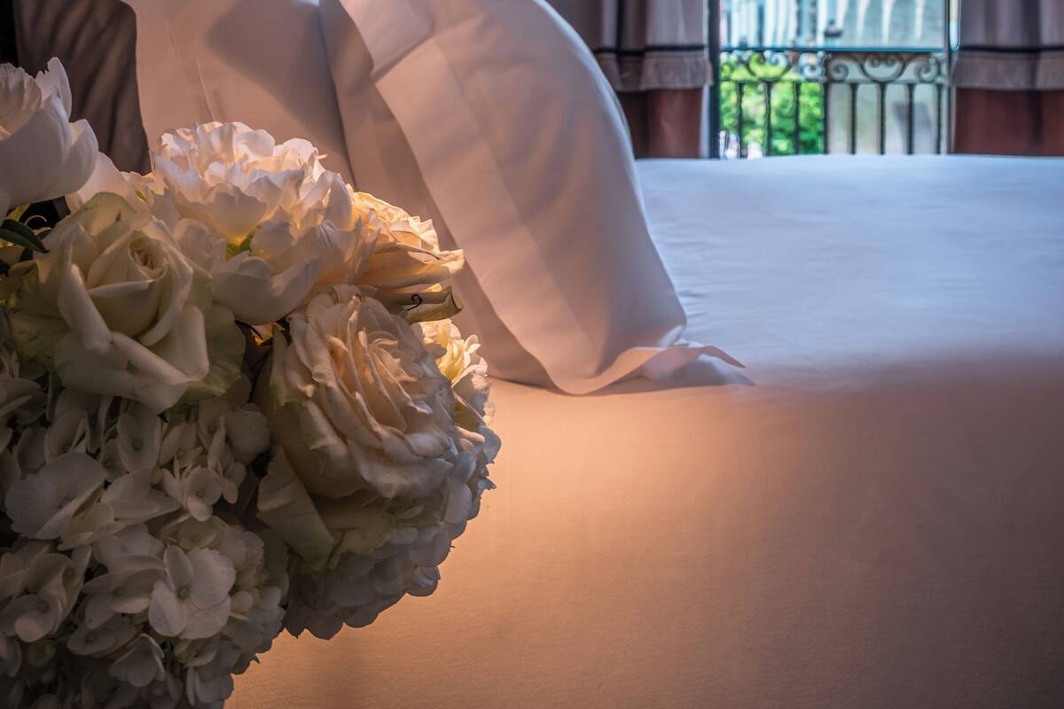 A hotel room with a bed, white pillows, a bedside lamp, a vase of flowers, and a view of a building through the window.