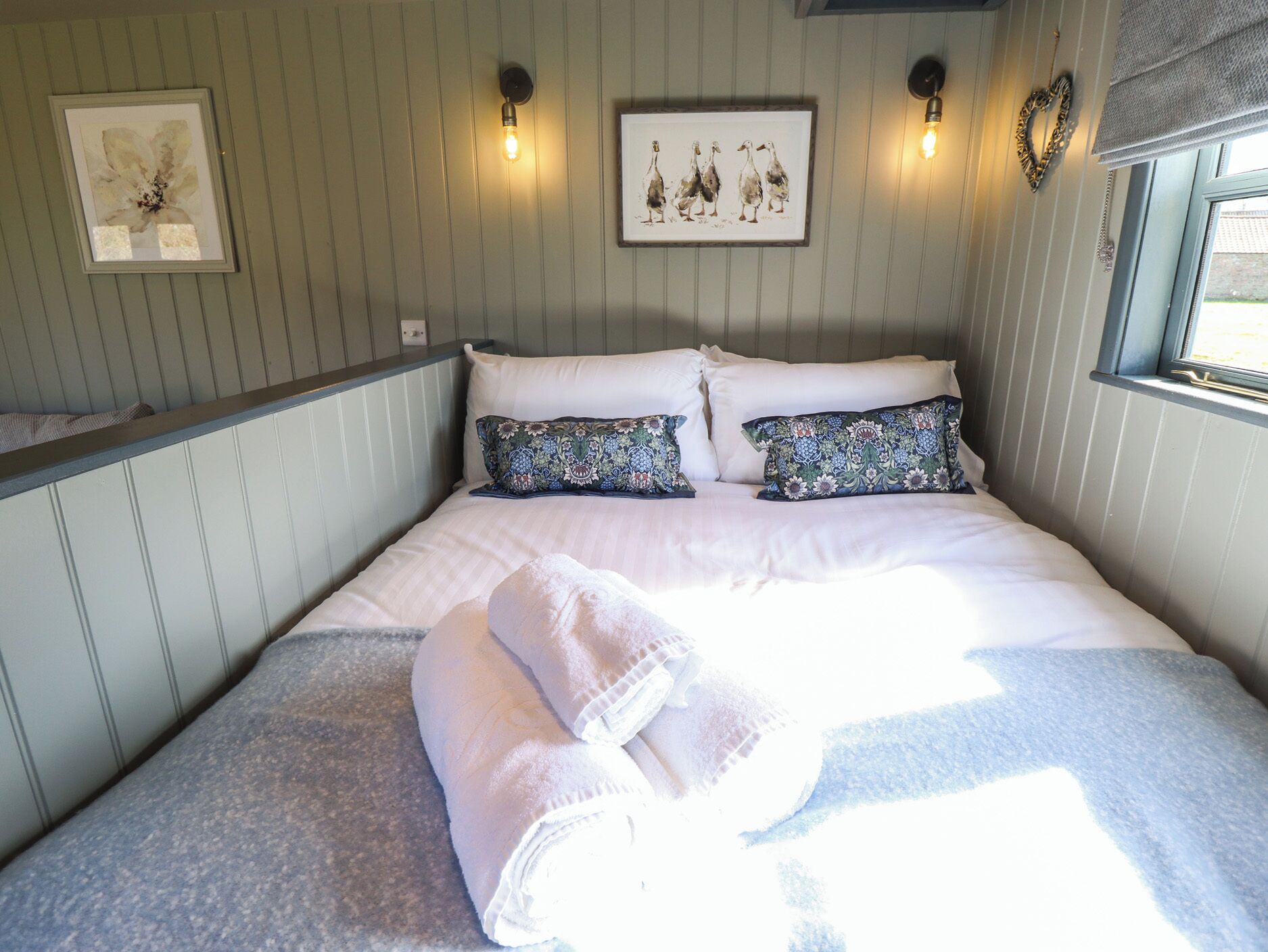A neatly made bed with white linens and decorative pillows, a framed artwork above the bed, and wall-mounted light fixtures.