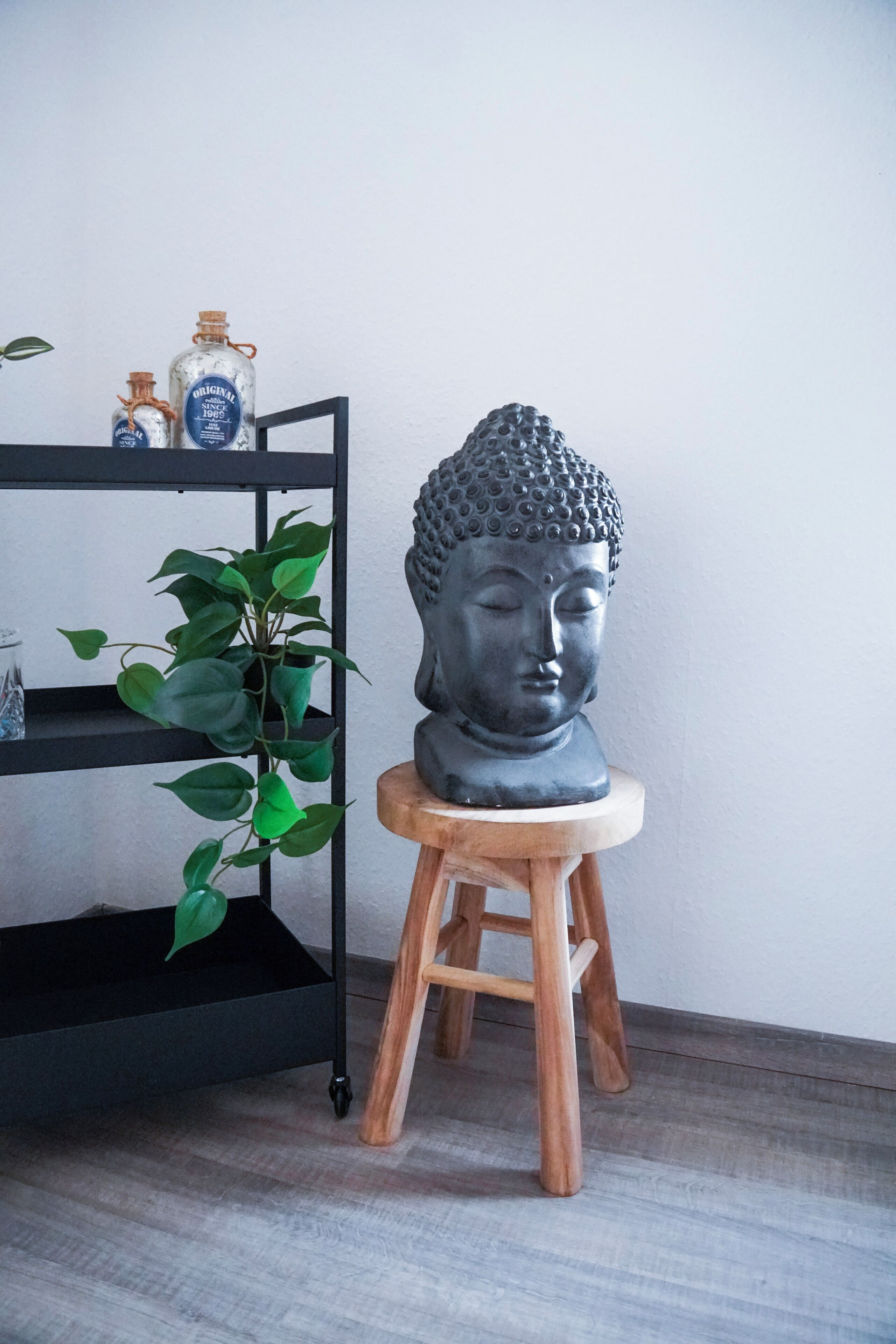 Une statue de Bouddha est posée sur un tabouret en bois, adossée à un mur blanc, à côté d’une étagère noire sur laquelle se trouvent deux bouteilles et une plante en pot.