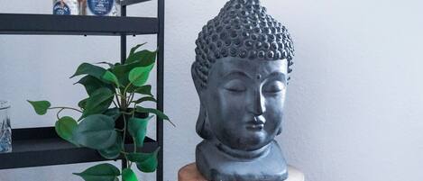Une statue de Bouddha est posée sur un tabouret en bois, adossée à un mur blanc, à côté d’une étagère noire sur laquelle se trouvent deux bouteilles et une plante en pot.