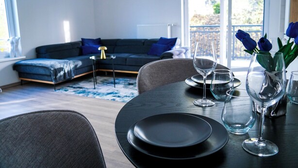 A modern living room with a dark-colored sofa, a dining table with plates and glasses, and a view of the outdoors through large windows.