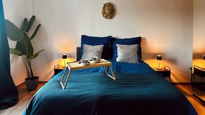 A bedroom with a large bed, two bedside tables with lamps, a chandelier, a potted plant, and a decorative wall piece.