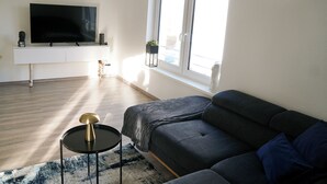 A modern living room with a large flat-screen TV, a dark-colored sofa, a small round table, and a window with a view of the outside.
