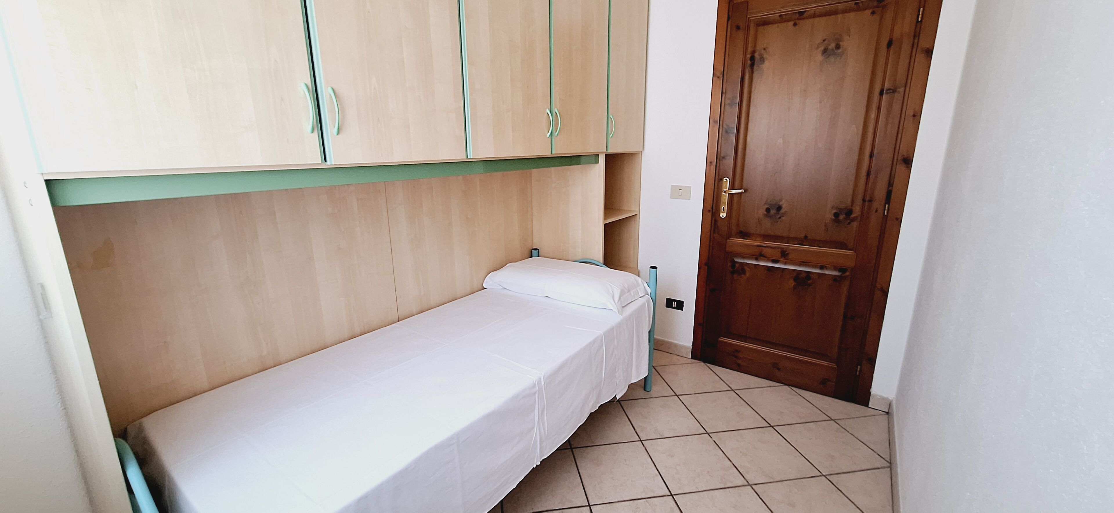 Ein kleines Zimmer mit einem Einzelbett, einem Holzschrank und einer Tür.