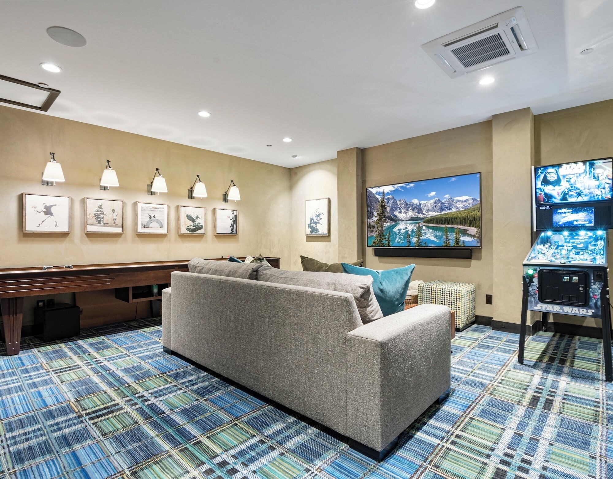 A room with a sofa, a pool table, wall-mounted lights, framed artwork, a large TV, and a Star Wars arcade machine.