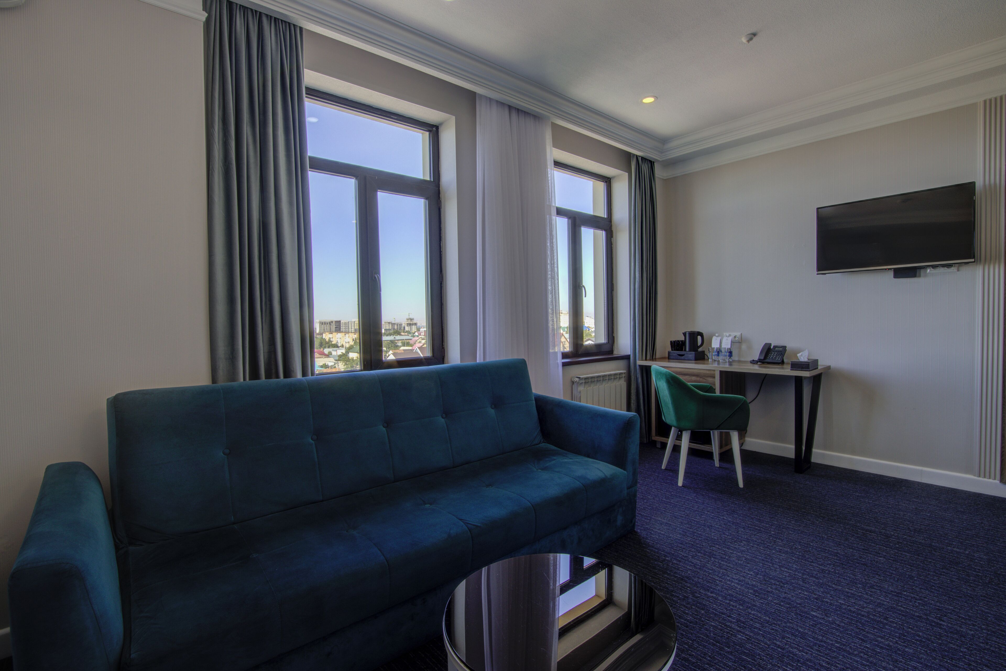 Habitación de hotel con sofá azul, un escritorio con silla, televisor montado en la pared y ventanas grandes con cortinas.
