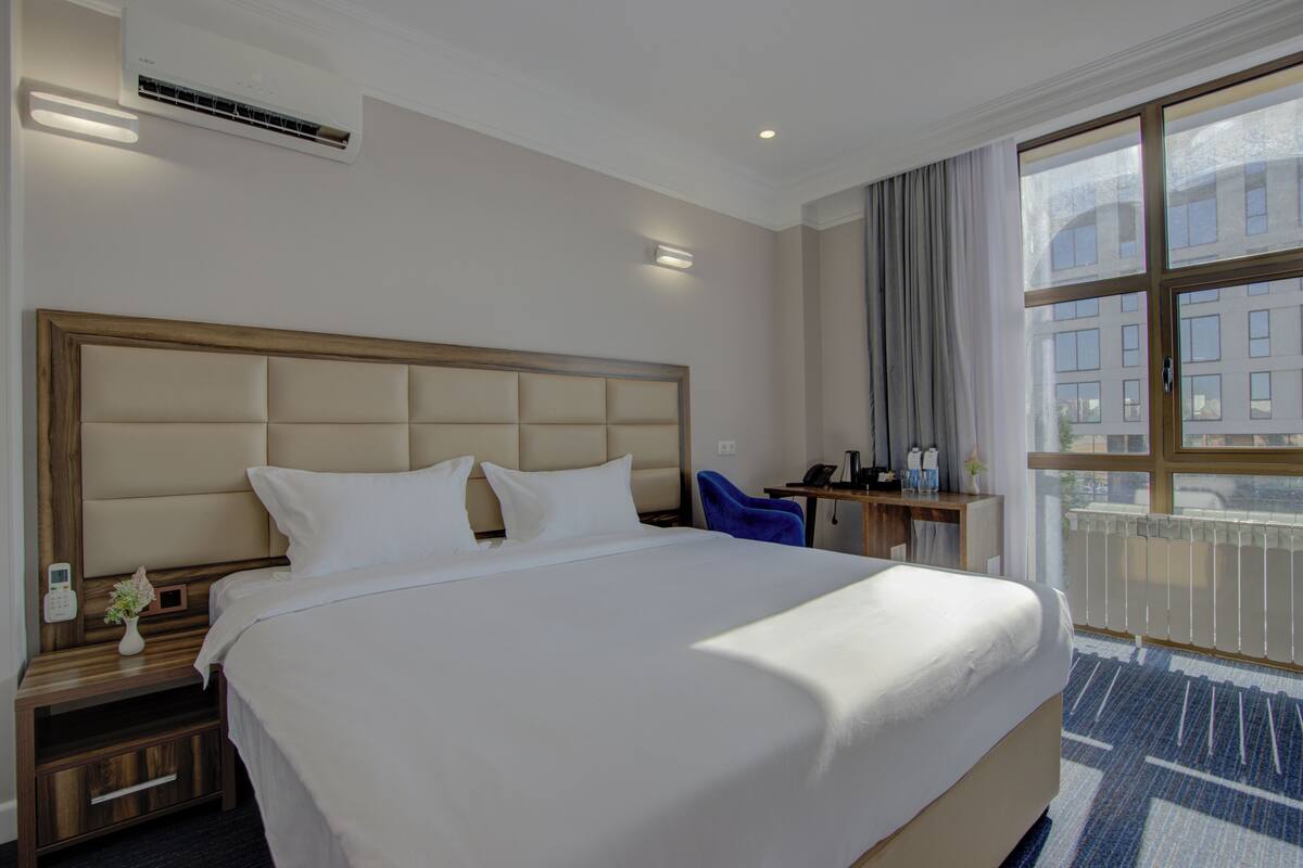 A hotel room with a large bed, a bedside table with a phone, a small table with a vase, and a window with a view of buildings.