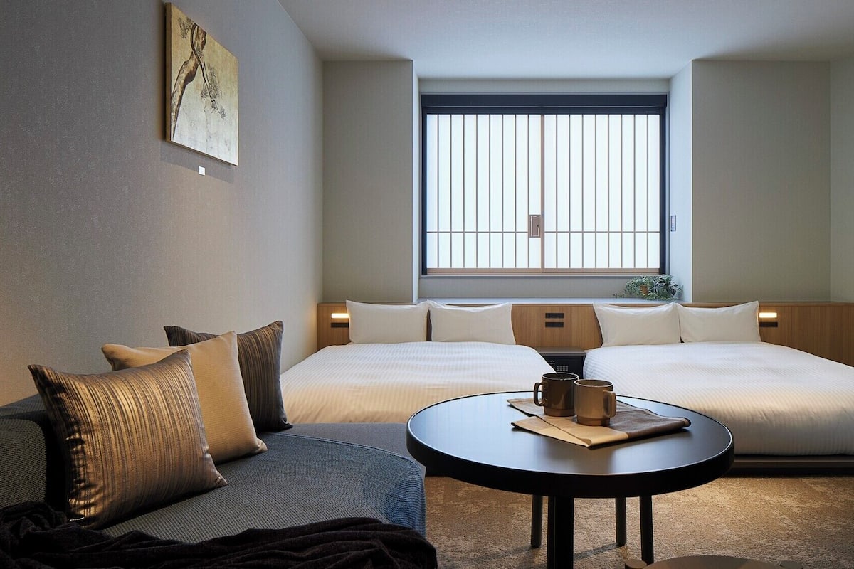 Habitación de hotel con una cama grande, un sofá, una pequeña mesa con una taza de café, un televisor y una ventana con persianas verticales.