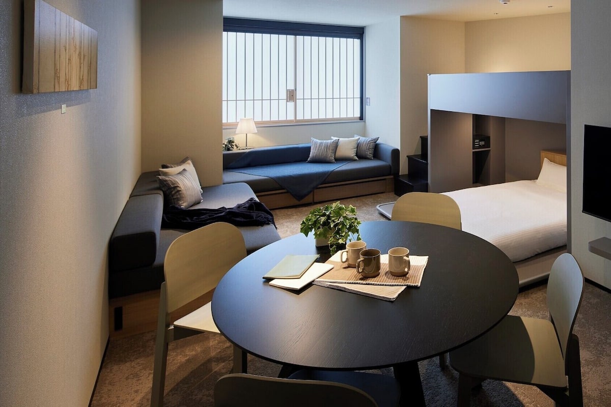 Habitación de hotel moderna con sofá, cama, televisor, mesa de comedor y ventana con persianas.