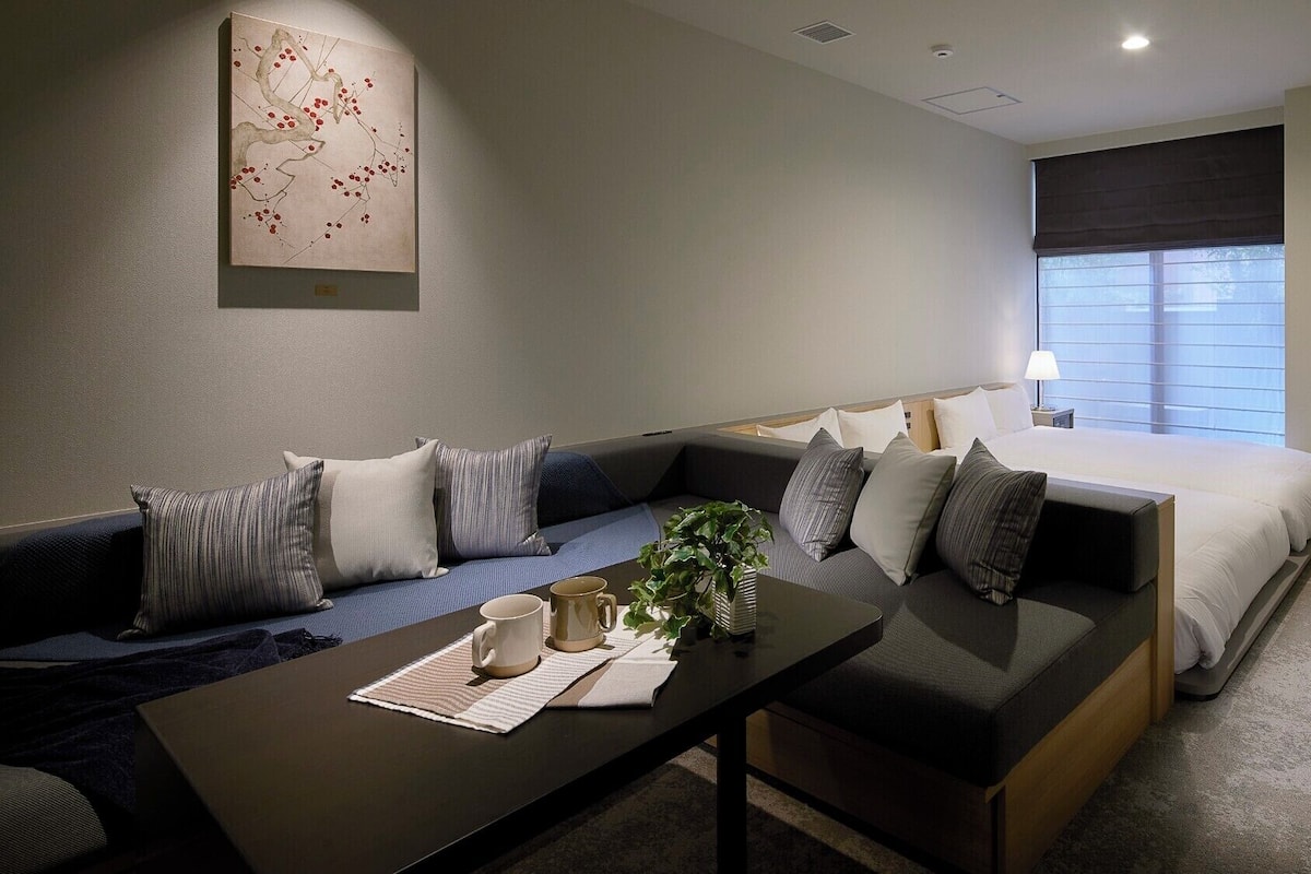 Una habitación de hotel moderna con un sofá, una cama, una mesita de noche con una planta y un ventanal grande.
