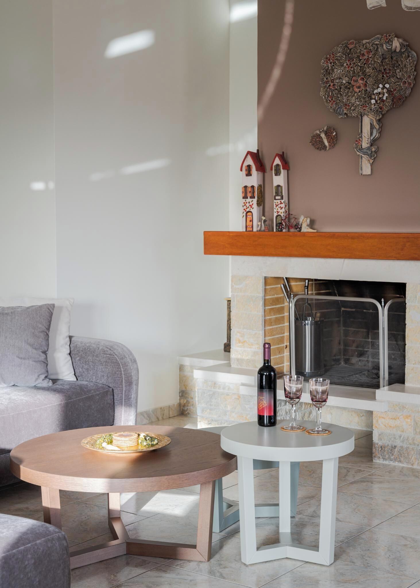 Una sala moderna con chimenea, un sofá gris, una mesa redonda de madera con una botella de vino y copas, y un adorno en la pared sobre la chimenea.