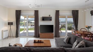 A modern living room with a large TV, a sofa, a coffee table, and a dining area with a view of the outdoors. - Ema's Guest House (Markopoulo Mesogaias)