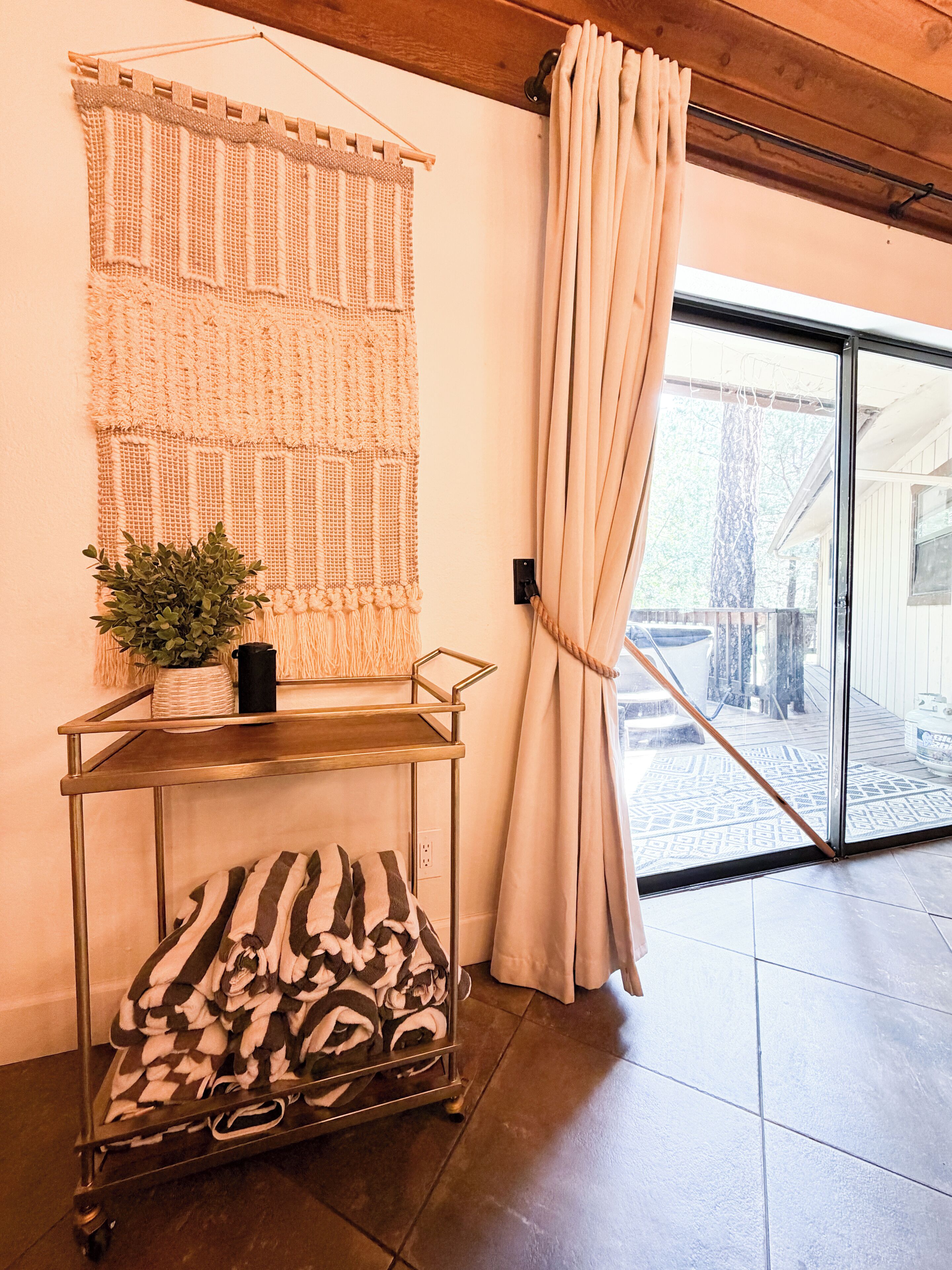 Una habitación con un tapiz tejido en la pared, un carrito metálico con toallas y vistas a través de una puerta de cristal.
