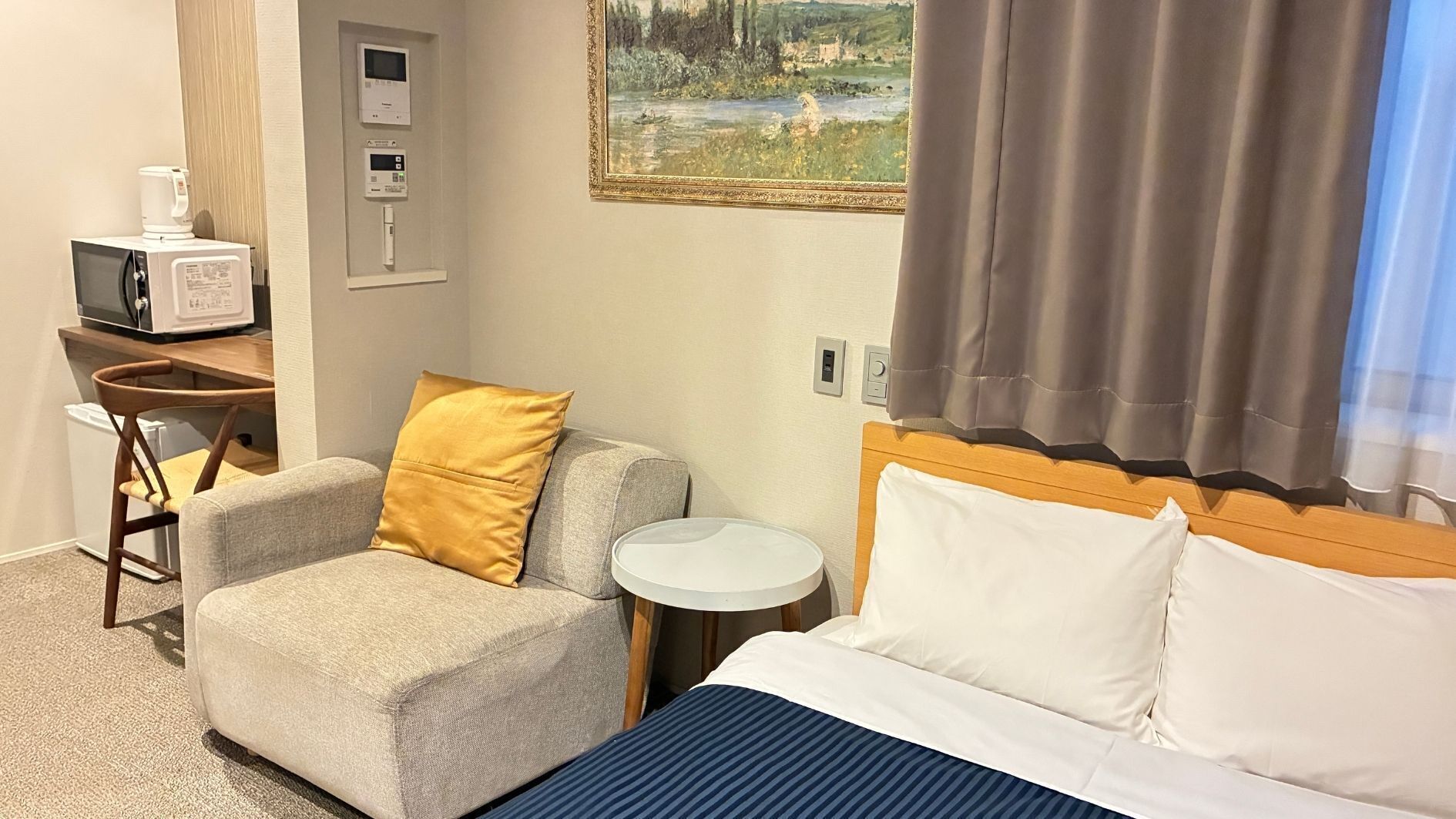 A hotel room with a bed, a chair with a pillow, a small table, a microwave, and a painting on the wall.