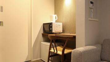 Een kleine, praktische kamer met een kitchenette, inclusief een magnetron en koffiezetapparaat. Er is een houten stoel en een bank, en een thermostaat aan de muur.
