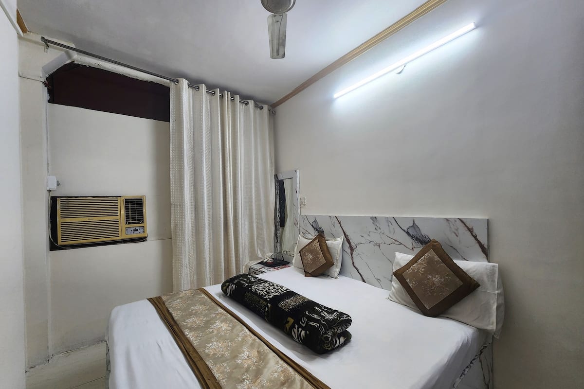 Una cama bien hecha con colcha blanca y dorada, dos almohadones decorativos, un calefactor en la pared y un ventilador de techo.