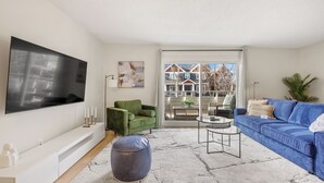A modern living room with a large flat-screen TV, green and blue sofas, a coffee table, and a view of a residential area through a glass door.