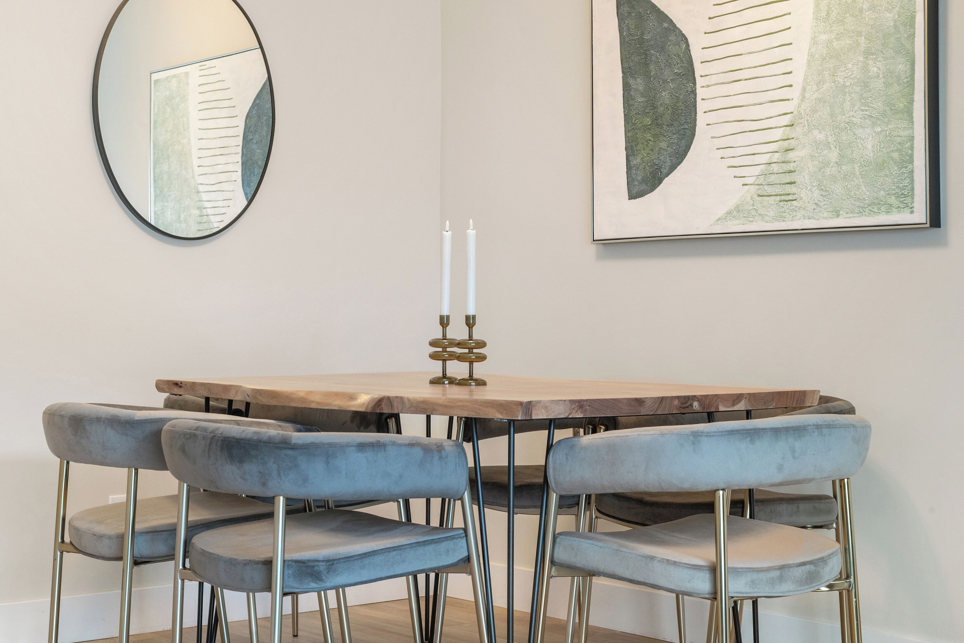 A dining area with a wooden table, four grey upholstered chairs, a mirror, and a framed abstract painting on the wall.
