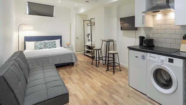 A compact hotel room with a bed, a sofa, a washing machine, a small kitchen area, and a mirror.