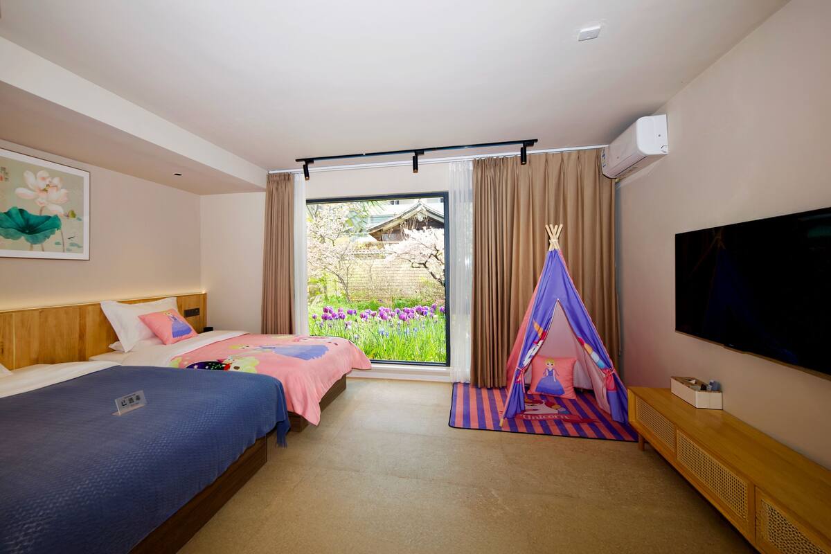 Ein Hotelzimmer mit zwei Betten, einem Fernseher und einem Fenster mit Blick auf Blumen und ein traditionelles Gebäude.