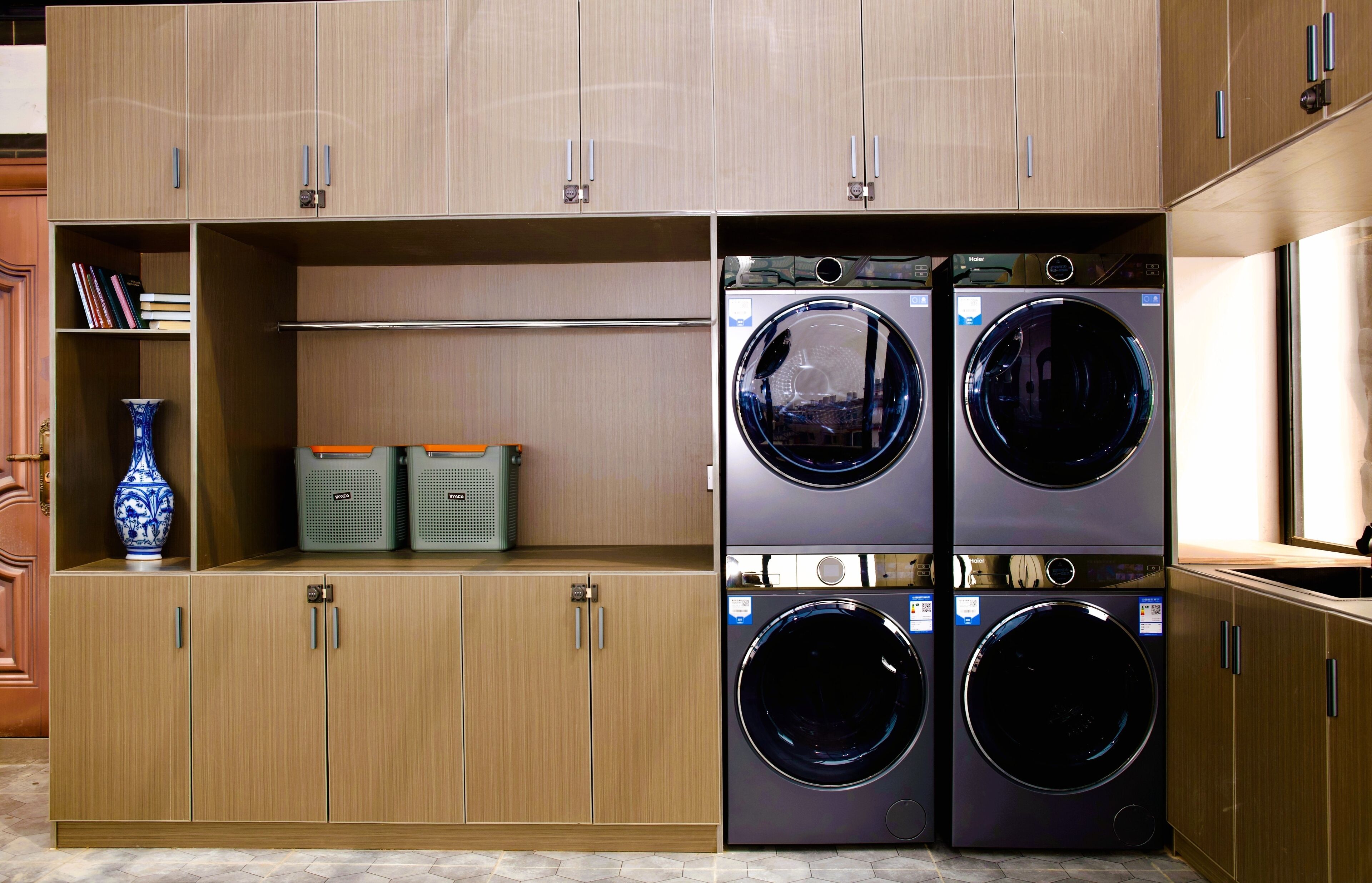 A laundry room with two stacked front-loading washing machines, wooden cabinets, and shelves with books and storage bins.