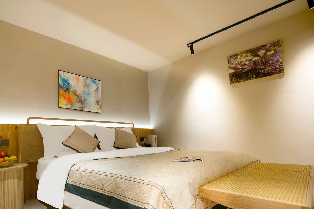 Ein modernes Hotelzimmer mit einem ordentlich bezogenen Bett, einem Nachttisch mit Früchten, einem kleinen Tisch mit einer Zeitschrift und einem Gemälde an der Wand.