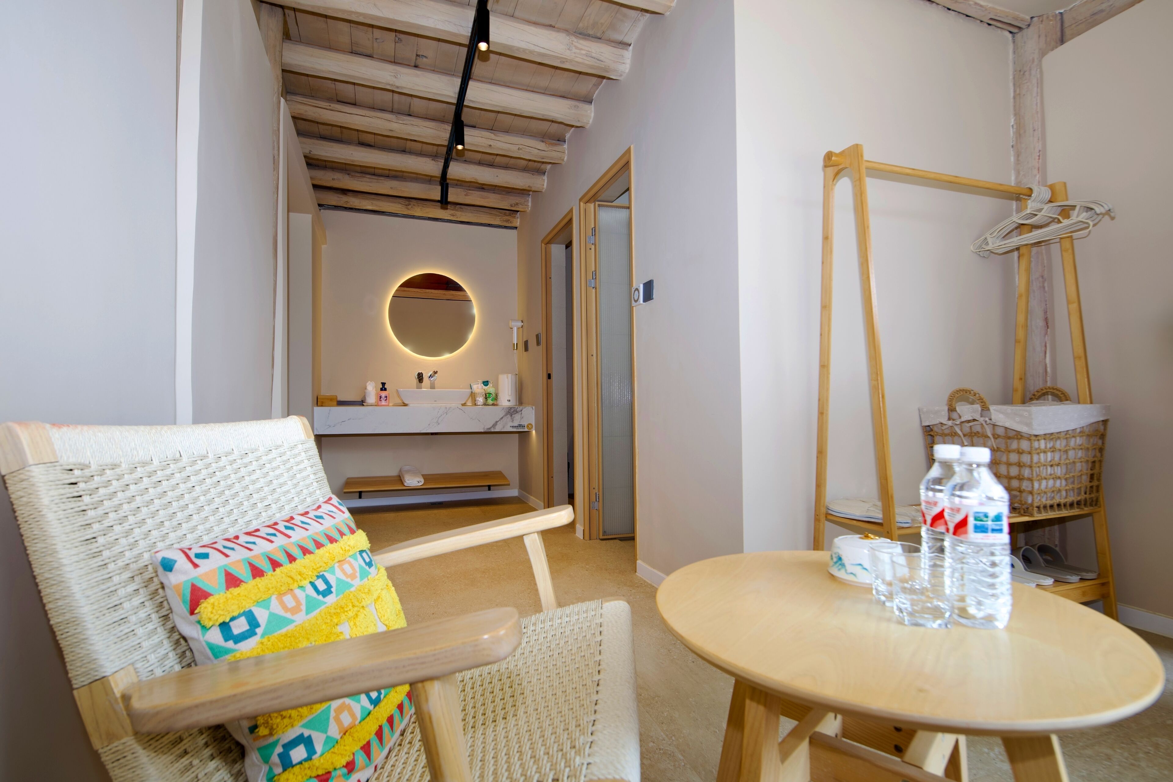 A room with a wicker chair, a round mirror, a wooden table with water bottles, and a wooden rack with shoes.