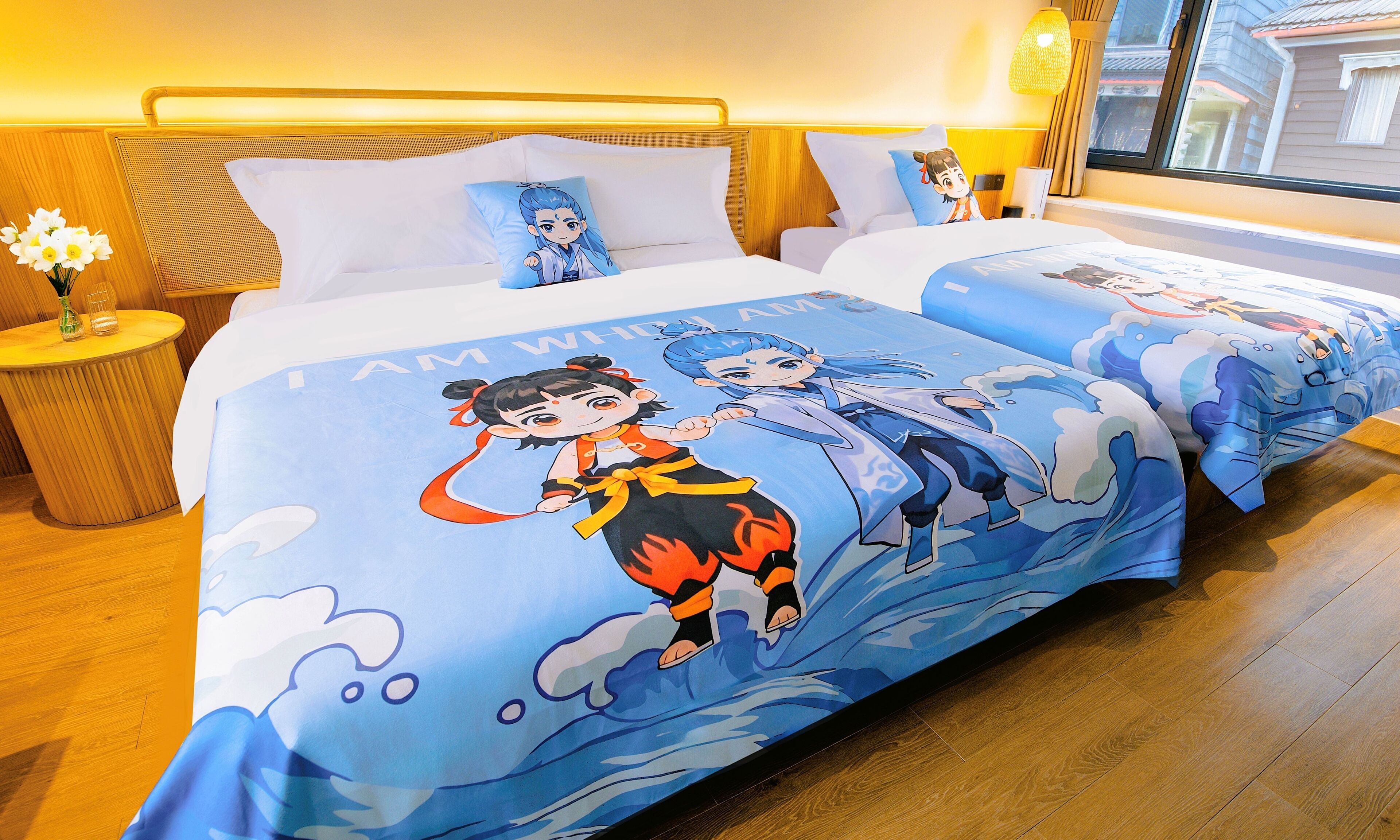 A hotel room with two beds, each with a blue bedspread featuring cartoon characters. There is a small wooden nightstand with a vase of flowers and a lamp.