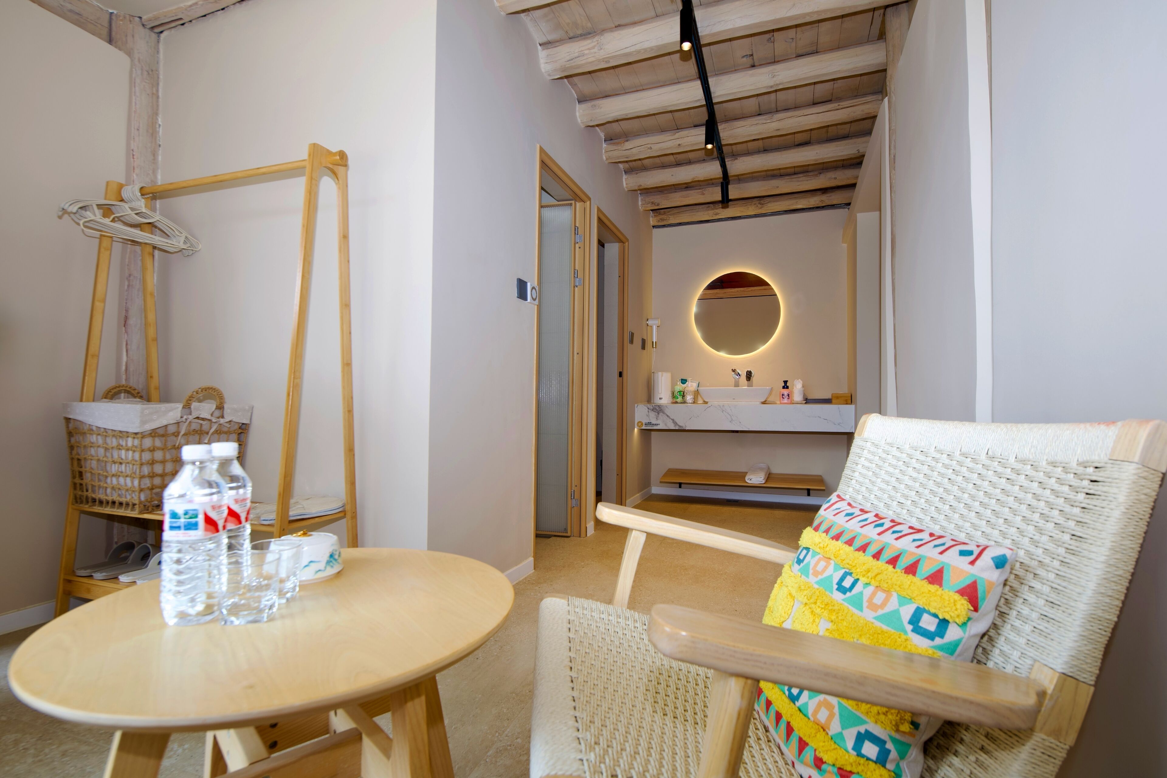 Una habitación con una mesa de madera, sillas de mimbre, una cesta y un espejo con luz redonda.