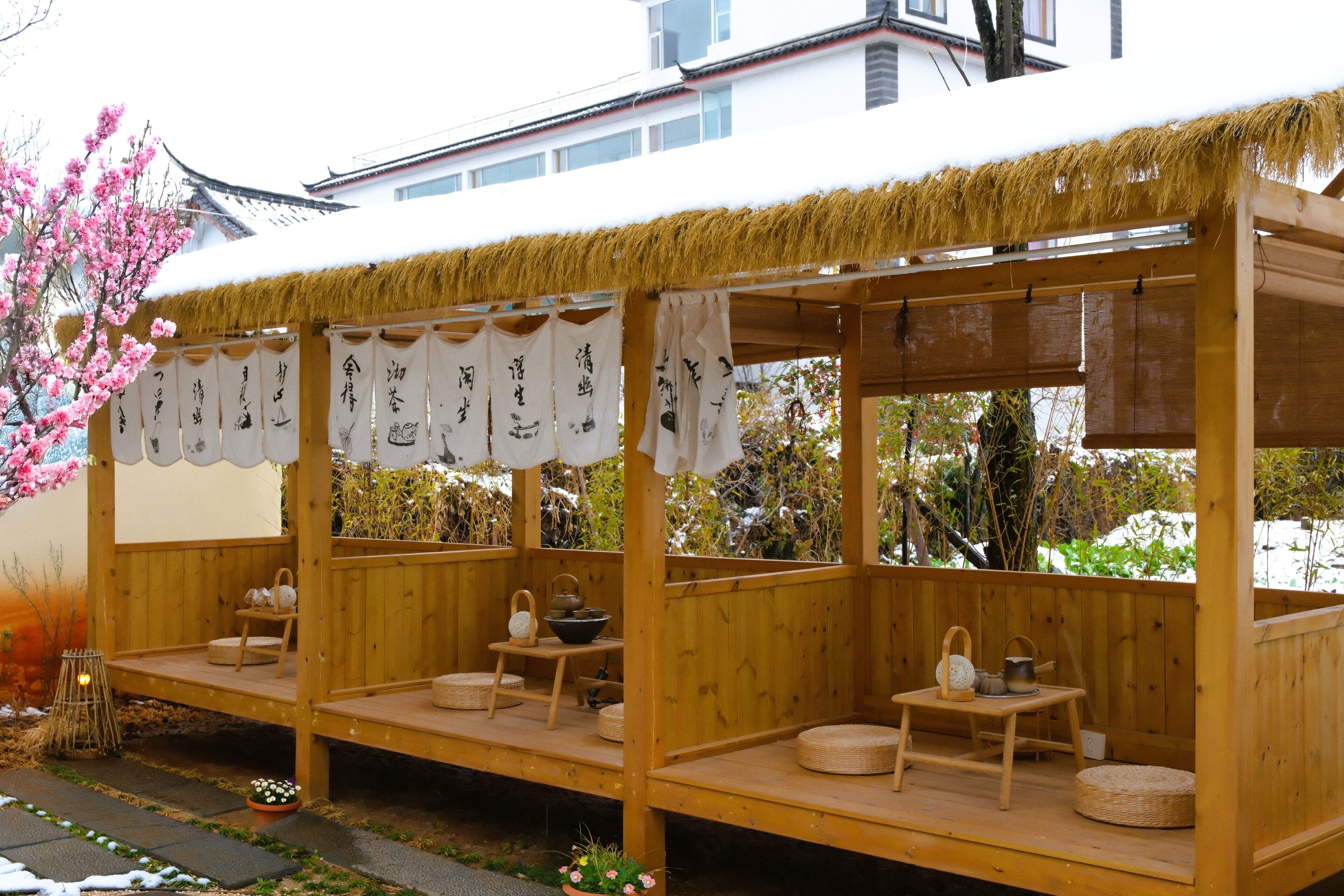 Eine traditionelle Holzkonstruktion mit Tatami-Matten und einem Strohdach, mit einem reetgedeckten Dach und Blick auf ein Gebäude im Hintergrund.
