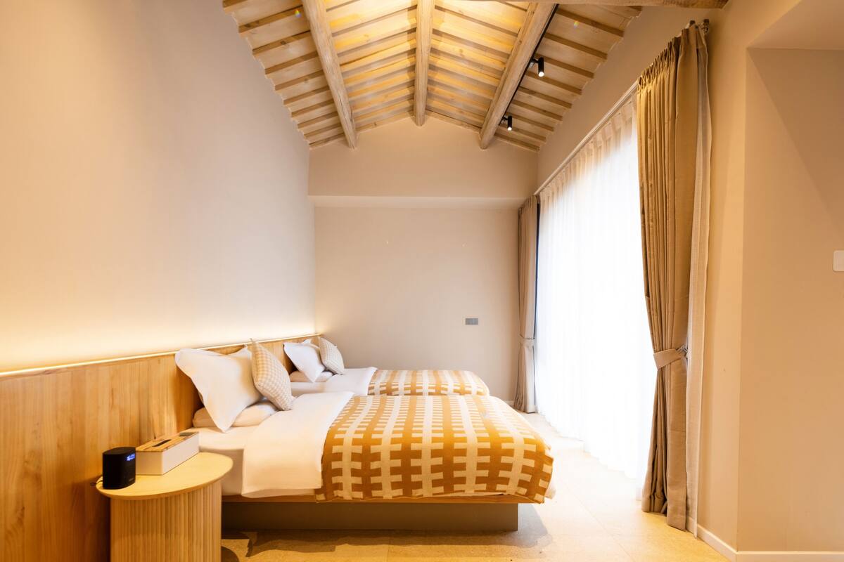 Ein Schlafzimmer mit Holzboden, einem Bett mit gemustertem Überwurf, einem Nachttisch mit Lampe und einem Fenster mit Vorhängen.