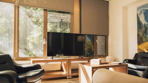 Ein modernes Wohnzimmer mit einem großen Flachbildfernseher, Holzmöbeln und großen Fenstern mit Blick ins Grüne.