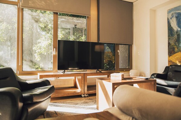 A modern living room with a large flat-screen TV, wooden furniture, and large windows with a view of greenery.