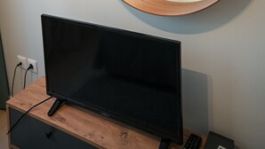 Ein Hotelzimmer mit rundem Spiegel, einem Bett mit weißer Bettwäsche, einem kleinen Nachttisch mit Lampe und einem Fernseher auf einem Holzständer.