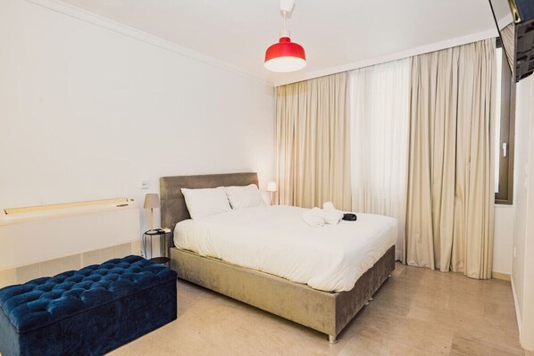 A hotel room with a bed, a bedside table, a lamp, a bench, and a red pendant light.