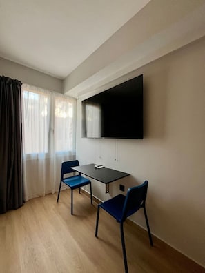 A room with a flat-screen TV mounted on the wall, a small table, two blue chairs, and a window with curtains. - Family Suite by Zeussuites (Athina)