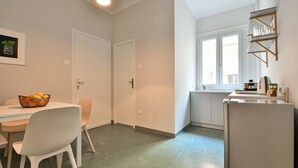 A compact kitchen with a dining area, white cabinetry, a refrigerator, a window, and a wall-mounted shelf. - Athenian Suite by Zeus suites (Athina)
