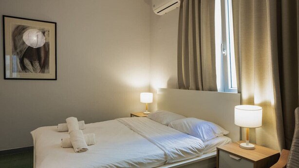 A hotel room with a bed, two bedside tables with lamps, a wall-mounted air conditioner, and a window with curtains. - Athenian Suite by Zeus suites (Athina)