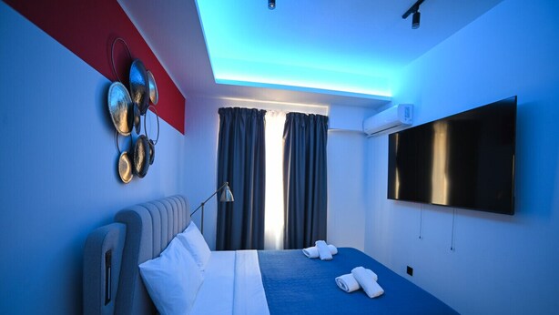 A modern hotel room with a bed, a flat-screen TV, and a ceiling light. - Muses Suite by Zeus suites (Athina)